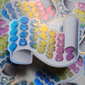 May include: A collection of colorful stickers featuring a high-heeled shoe design. Each shoe is adorned with rows of small, smiling frog illustrations in a rainbow of colors, including blue, yellow, pink, and green. The stickers have a holographic, iridescent finish.