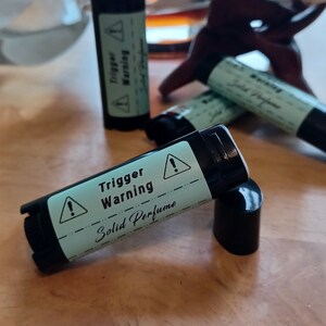 May include: Black solid perfume tubes with a light green label that reads "Trigger Warning" and "Solid Perfume". The label also features an exclamation point warning symbol. The tubes are on a wooden surface.