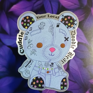May include: A light blue sticker featuring a cartoon bear with a stitched design. The bear has brown eyes, a pink nose, and various tattoos. The sticker includes the text "Cuddle Doodle Bear" and "Your Local".