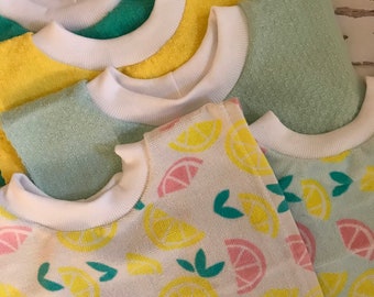 May include: A stack of baby bibs in various pastel colors, including yellow, green, and blue. The bibs have a white ribbed collar and are decorated with a lemon pattern. The bibs are made of a soft, absorbent material.