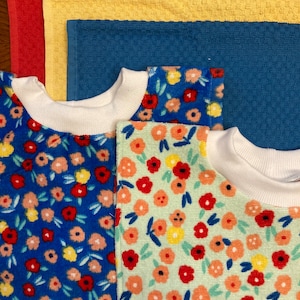 May include: An overhead shot of colorful towels and two floral print shirts. The towels are red, yellow, and blue. The shirts have white collars and are patterned with red, yellow, and orange flowers on blue and mint green backgrounds.