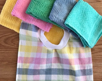 May include: A baby bib with a pastel plaid pattern, featuring a white collar and a yellow backing. Five small washcloths in yellow, pink, green, blue, and teal are arranged above the bib. The items are made of a soft, textured fabric.
