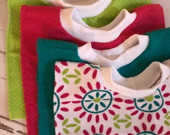 May include: A stack of colorful, folded terry cloth shirts. The shirts are in shades of green, red, and teal, with white collars. One shirt features a white background with a floral pattern in red, green, and teal.