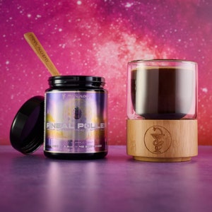 Puede incluir: Un frasco negro con una etiqueta morada y dorada que dice "Pineal Pollen Aura Enhanced Mind & Spirit Elixir" con una cuchara de madera encima. El frasco está sobre una superficie morada junto a una taza de vidrio con un líquido oscuro dentro y una base de madera con un símbolo de serpiente y bastón.