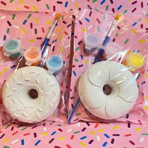 May include: Two paint-your-own donut kits in clear plastic bags. Each kit includes a white ceramic donut, paint pots in various colours, and a paintbrush. The donuts have a raised design and are ready to be painted. The background is pink with colourful sprinkles.