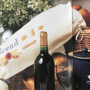 Peut inclure: Un sac à pain en lin blanc avec du blé brodé et le mot "Pain". Une bouteille de vin rouge, un verre de vin rouge et un panier en osier avec du pain sont également présents sur l'image.