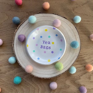 May include: A white ceramic dish with colourful polka dots and the words "Tea Bags" in purple lettering. The dish is surrounded by a variety of colourful felt balls.