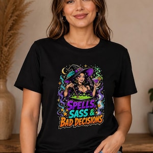 May include: A black short-sleeve t-shirt featuring a colorful graphic of a witch stirring a green cauldron while holding a cocktail. The design includes the text "SPELLS SASS & BAD DECISIONS" in bold, stylized lettering surrounded by stars and smoke.