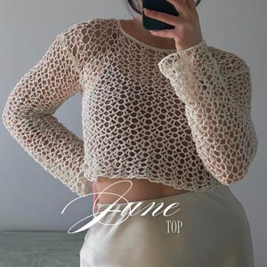 Puede incluir: Una blusa de manga larga tejida a ganchillo en color crema con un patrón de malla abierta, cuello redondo y dobladillo corto. La prenda se lleva sobre una prenda interior oscura e incluye el texto cursivo blanco Jane TOP en la imagen.