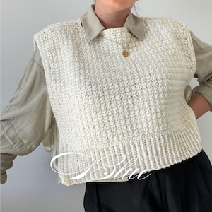 Puede incluir: Un chaleco de ganchillo sin mangas de color crema con un patrón de punto texturizado y un dobladillo acanalado. La prenda se lleva sobre una camisa de cuello verde oliva claro y pantalones negros. El texto "iBlue" se ve en letra blanca.