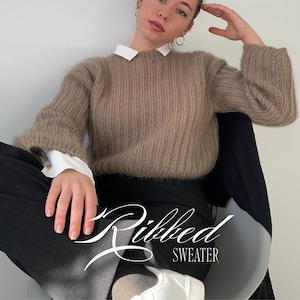 Puede incluir: Un suéter de manga larga acanalado en color topo con cuello redondo, usado sobre una camisa blanca con cuello. El suéter presenta una textura difusa y canalé vertical. El texto "Ribbed SWEATER" está superpuesto en letras blancas.