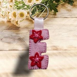 Könnte beinhalten: Ein handgefertigter Schlüsselanhänger mit dem Buchstaben "E" aus rosa Filz, verziert mit zwei roten Blumenapplikationen. Der Anhänger hat einen silbernen Ring und eine weiße Schleife. Hintergrund: Holzoberfläche und weiße Blumen.