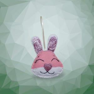 Könnte beinhalten: Ein rosa Filz-Hasen-Ornament mit glitzernden Ohren und einer herzförmigen Nase. Der Hase hat ein lächelndes Gesicht mit schwarzen gestickten Details und einer weißen Kontur. Ein cremefarbenes Band ist zum Aufhängen befestigt.