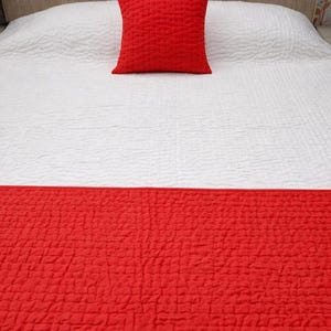 May include: A bed with a white textured quilt and a bright red quilted pillow. A matching red bed runner is at the foot of the bed, creating a bold contrast. The bedding is neatly arranged, suggesting a comfortable and stylish bedroom.