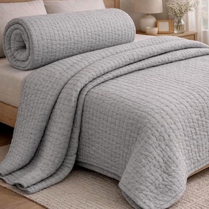 Può includere: Un set di copriletto e cuscino a rullo trapuntati grigio chiaro. Il copriletto è drappeggiato su un letto, con il cuscino a rullo arrotolato alla testata. La biancheria da letto presenta un motivo a griglia strutturato.