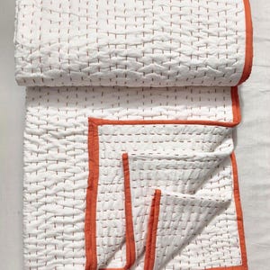 May include: White quilted blanket with orange trim. The blanket features a repeating pattern of small, evenly spaced stitches in a contrasting color. The edges are finished with a bright orange border, adding a pop of color to the otherwise neutral design.