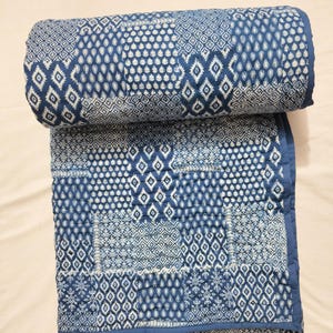 Könnte beinhalten: Eine gerollte und teilweise entfaltete blau-weiß gemusterte Steppdecke. Die Steppdecke weist ein sich wiederholendes geometrisches Design mit Diamanten- und Tropfenformen auf. Die Kanten sind mit einem durchgehenden blauen Rand versehen. Die Steppdecke besteht aus einem weichen, gesteppten Material.