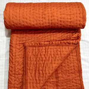 May include: An orange quilted blanket, folded and rolled, with a textured surface. The blanket is a solid colour and appears to be made of a soft material. The stitching creates a grid-like pattern across the surface.