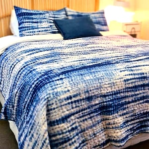 May include: A bed with a woven rattan headboard and bedding featuring a blue and white tie-dye pattern. The bed includes matching pillowcases and a solid blue decorative pillow. The overall aesthetic is coastal and relaxed.