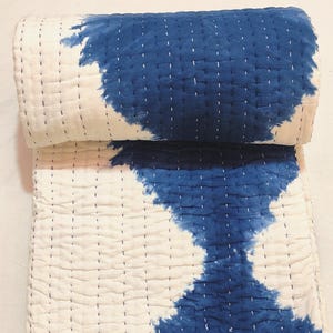 May include: A rolled-up, white and blue quilt with a tie-dye pattern. The quilt is made of a soft material and features a stitched design. The blue dye creates an abstract design on the white background. The quilt is folded over on itself.