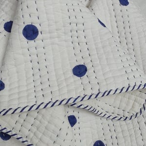 May include: White quilted blanket with navy blue polka dots and navy blue striped trim. The blanket has a stitched grid pattern and is folded over. The blanket is made of a soft, textured material.