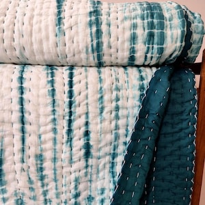 May include: White and teal tie-dye patterned quilt. The quilt features a vertical stripe design with teal dye on a white background. The quilt is made of a soft material and has white stitching.