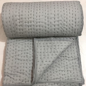 May include: A light grey quilt with black stitching. The quilt is folded and rolled, displaying the detailed stitching pattern. The fabric appears soft and textured, suitable for bedding or home decor.