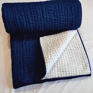 May include: A folded quilt featuring a deep navy blue top side and a contrasting white underside. Both sides of the textile display a consistent grid-like pattern of small, straight running stitches in a dark thread, creating a textured appearance.