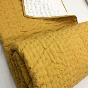 Könnte beinhalten: Eine gefaltete, senfgelbe Steppdecke mit weißem Rand. Die Steppdecke weist ein genähtes, gitterartiges Muster auf. Der weiße Rand ist an der Oberkante mit gelbem Besatz sichtbar. Die Steppdecke ist gefaltet und zeigt die Textur und das Design.