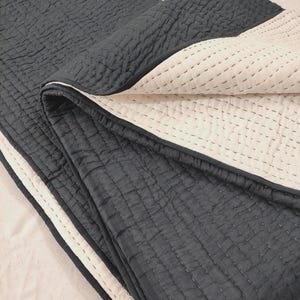 May include: A folded, reversible quilt with a dark gray top layer and a cream-colored underside. The quilt features a stitched, grid-like pattern across both sides. A white border with a dark gray trim is visible along the edge.