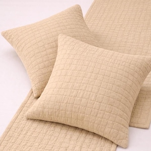 May include: Two beige square pillows and a matching bed runner. The pillows and runner have a quilted grid pattern. The items are made of a soft, textured fabric, and are arranged on a white surface.