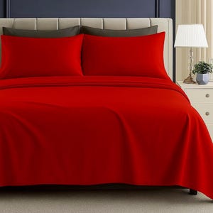 May include: A bed is made with a bright red sheet set. Two red pillowcases are visible, resting on dark gray pillowcases. The bed is against a neutral-colored headboard. A nightstand with a lamp and a small framed picture is to the right.