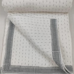 Pode incluir: Colcha branca com um padrão repetido de traços cinzentos verticais curtos. A colcha tem uma borda de listras cinzentas e é dobrada, revelando o interior. A colcha é feita de um tecido macio e texturizado.