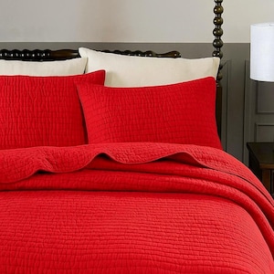 May include: A red quilted bedspread and pillow set. The bedding has a textured, brick-like pattern. The pillows are rectangular and the bedspread is neatly arranged on the bed.