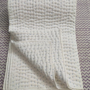 May include: A white cotton quilt with blue stitching, folded in half and lying on a purple and white patterned surface.