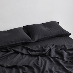 Puede incluir: Juego de ropa de cama de lino de color carbón oscuro, que incluye dos fundas de almohada y una sábana bajera. La tela está ligeramente arrugada, lo que sugiere una textura suave y cómoda. La ropa de cama está colocada sobre una cama, con un fondo neutro.