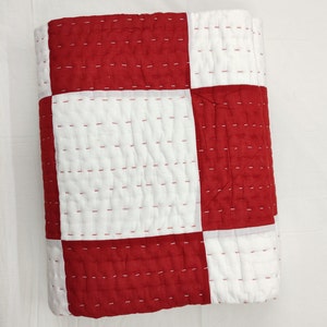 Op de afbeelding: Gevouwen rood-witte patchworkdeken. De deken heeft een dambordpatroon met grote vierkanten van effen rode en witte stof. De deken is gestikt met rode draad.