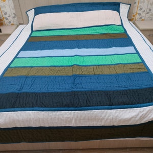 May include: A striped quilt in shades of blue, green, and white, with a solid blue border. The quilt is laid out on a bed, showcasing its design. The quilt's dimensions are not visible in the image.