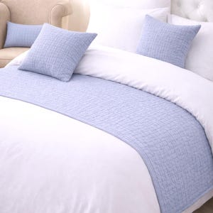 May include: A bed with white bedding, featuring a light blue quilted bed runner and matching square pillows. A smaller rectangular pillow is visible on a beige armchair in the background. The pillows and runner have a textured pattern.