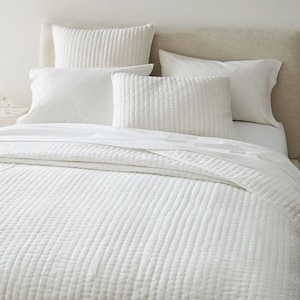 May include: A bed dressed in white bedding. It features a quilted duvet cover, multiple pillows, and a flat sheet. The pillows vary in size and shape, some with a quilted design. The headboard is a neutral tone.