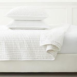 May include: A white bed set comprising a quilted blanket and two pillows. The blanket and top pillow have a textured, stitched pattern. The bed set is on a white bed frame, creating a clean, minimalist aesthetic.