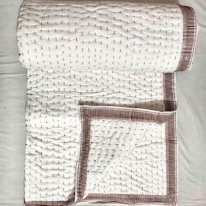 Op de afbeelding: Een witte gewatteerde deken met een bruine rand en stiksels. De deken is gedeeltelijk opgerold, waardoor de binnenkant zichtbaar is. De stiksels zijn gelijkmatig verdeeld over het oppervlak, waardoor een textuur ontstaat. De rand is effen bruin.
