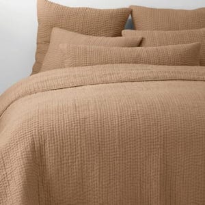 Könnte beinhalten: Ein hellbraunes Bettwäsche-Set mit einer Steppdecke und mehreren passenden Kissen. Die Bettwäsche hat ein strukturiertes, gestepptes Design. Die Kissen sind in verschiedenen Größen und die Steppdecke bedeckt das Bett. Der Hintergrund ist eine schlichte weiße Wand.