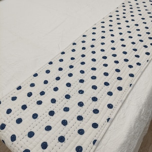 Puede incluir: Un camino de cama blanco con un patrón repetido de lunares azul marino. El camino está acolchado con costuras visibles y se coloca sobre una colcha blanca. El diseño es limpio y moderno, adecuado para la decoración del dormitorio.