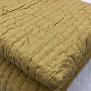 Könnte beinhalten: Senffarbene Steppdecke mit weißen Nähten. Die Decke ist gefaltet und zeigt die Textur des Stoffes und das Steppmuster. Das Gesamtbild ist weich und einladend, geeignet für ein Bett oder Sofa.