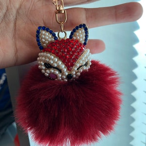 Op de afbeelding: Een rode, pluche sleutelhanger met een bejeweled vos-bedel. De vos is versierd met rode, witte en blauwe strass steentjes en parels.