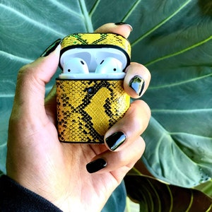 Pode incluir: Uma caixa para fones de ouvido sem fio com padrão de pele de cobra amarela e preta. A caixa está aberta e os fones de ouvido brancos são visíveis no interior.