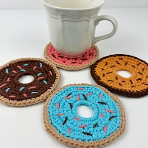 May include: A set of handmade crochet donut coasters in various colours. The coasters feature a donut design with sprinkles, including brown, blue, and orange. A white mug sits on a pink coaster. The coasters are designed to protect surfaces from heat and spills.