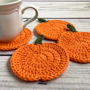May include: Orange crochet pumpkin coasters with green stems. A white mug sits on one coaster. The handmade coasters are on a light wood surface. These decorative coasters are perfect for autumn decor.