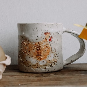 Könnte beinhalten: Eine weiße Keramiktasse mit einem braunen und gelben Hühnerdesign. Die Tasse steht auf einem Holztisch neben einer Eierpackung und einer kleinen braunen Vase mit gelben Narzissen.